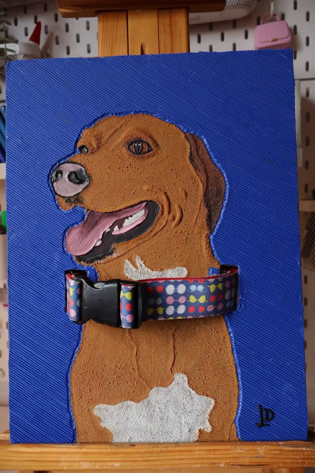 Cuadro en plastilina y en relieve de un perro café sobre fondo azul, hecho en su memoria, con su collar real incorporado a la obra