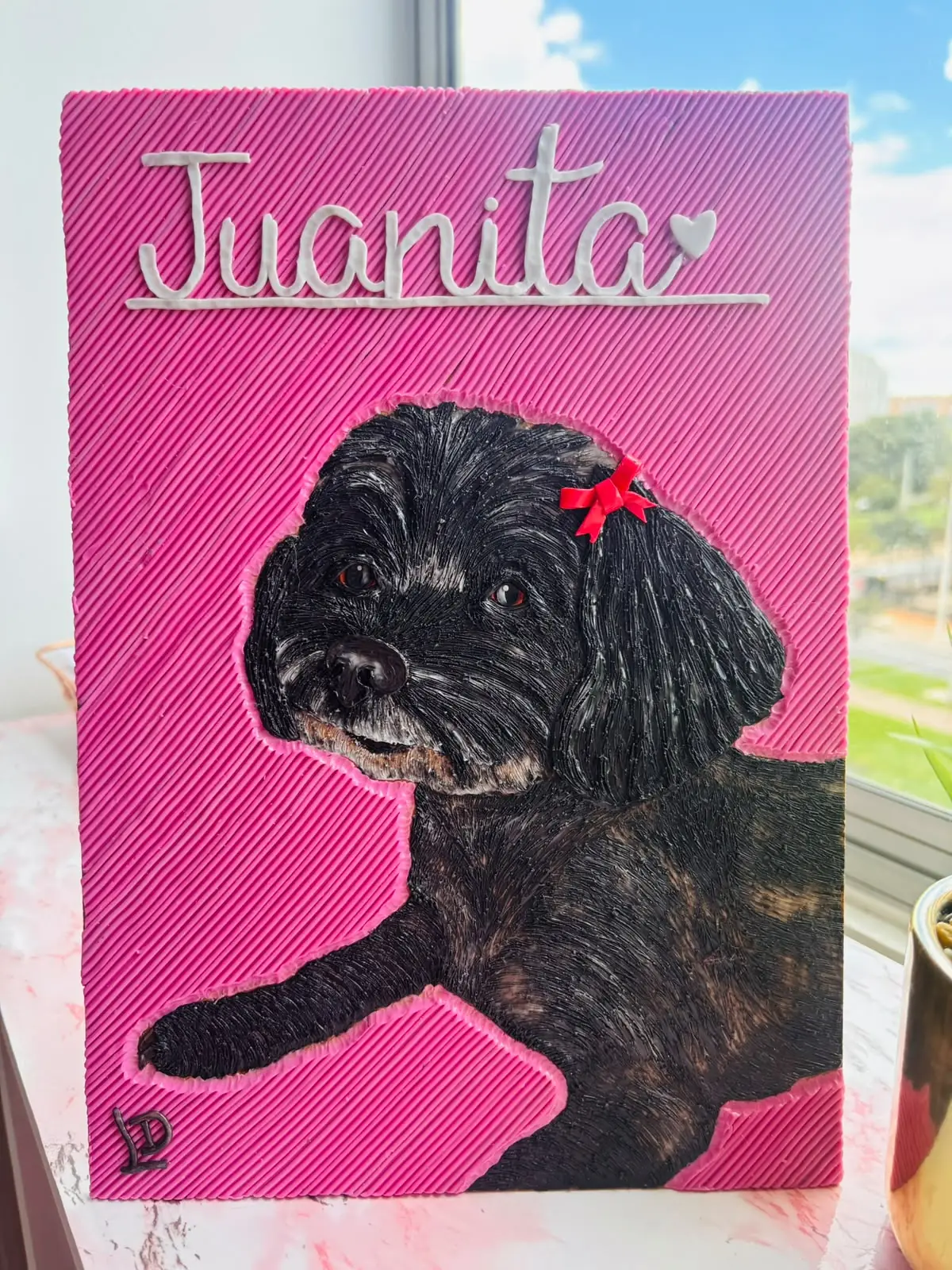 Cuadro en plastilina y en relieve de Juanita, una perrita negra con un moño rojo, sobre fondo fucsia